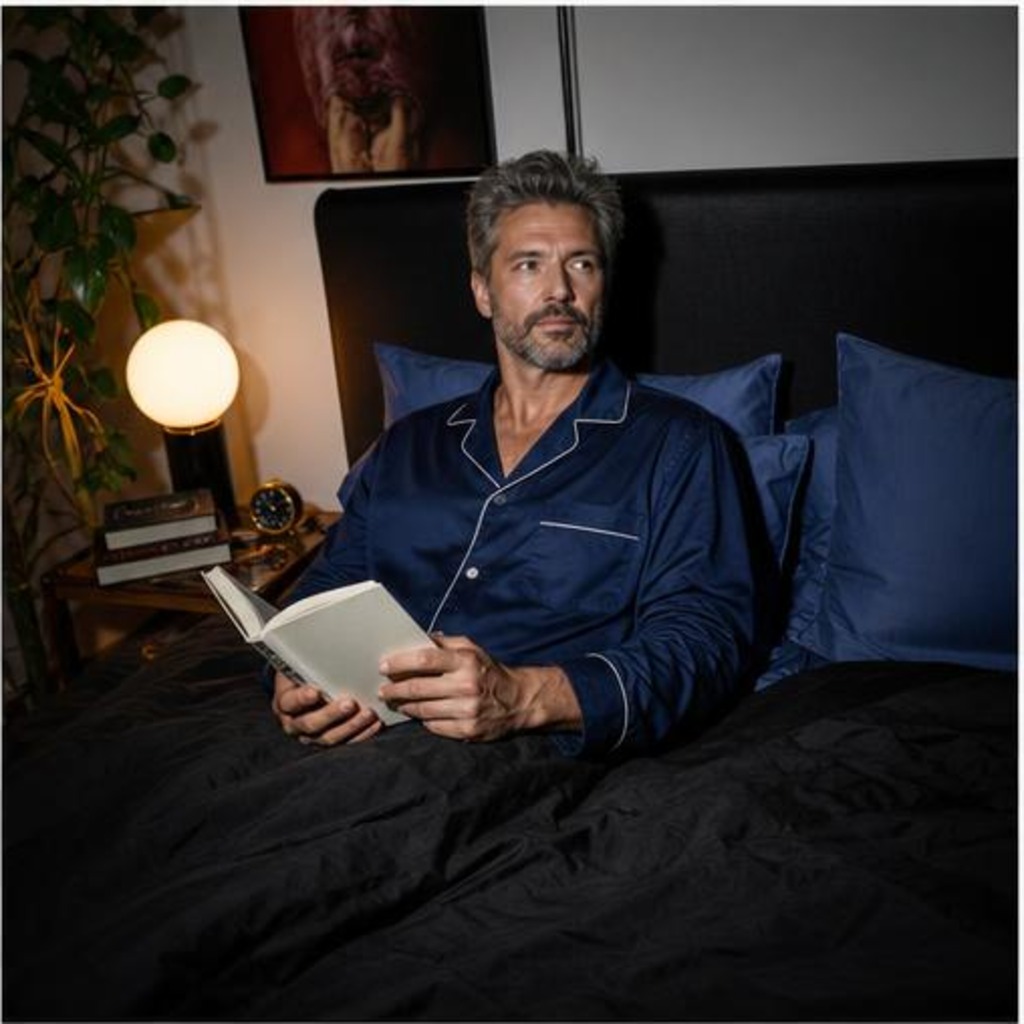 Fictional man in navy pajamas holding an open book in bed beside blue pillows and a warm bedside lamp.