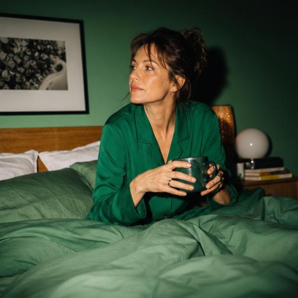 Fictional woman in green sleepwear sitting in bed with green bedding, holding a mug beside a bedside lamp.