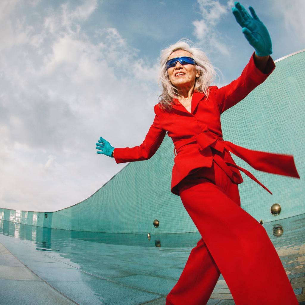 Silver-haired woman in a red suit dancing in a turquoise tiled plaza.