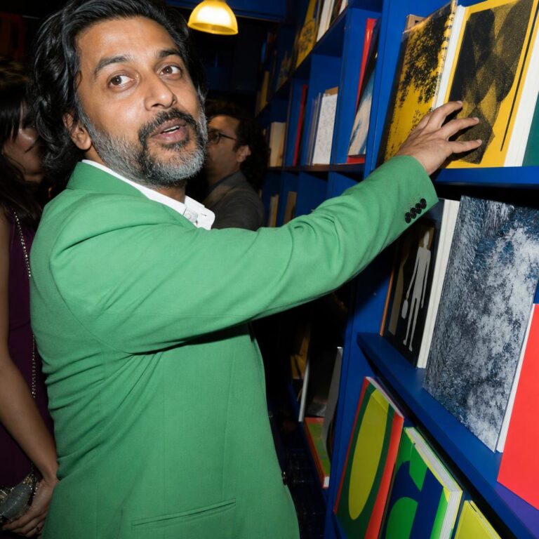 South Asian man in a green suit reaching for an art book in a blue bookshop.