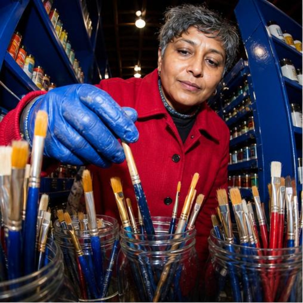 Fictional woman in a red coat selecting paintbrushes in a cobalt art-supply shop.