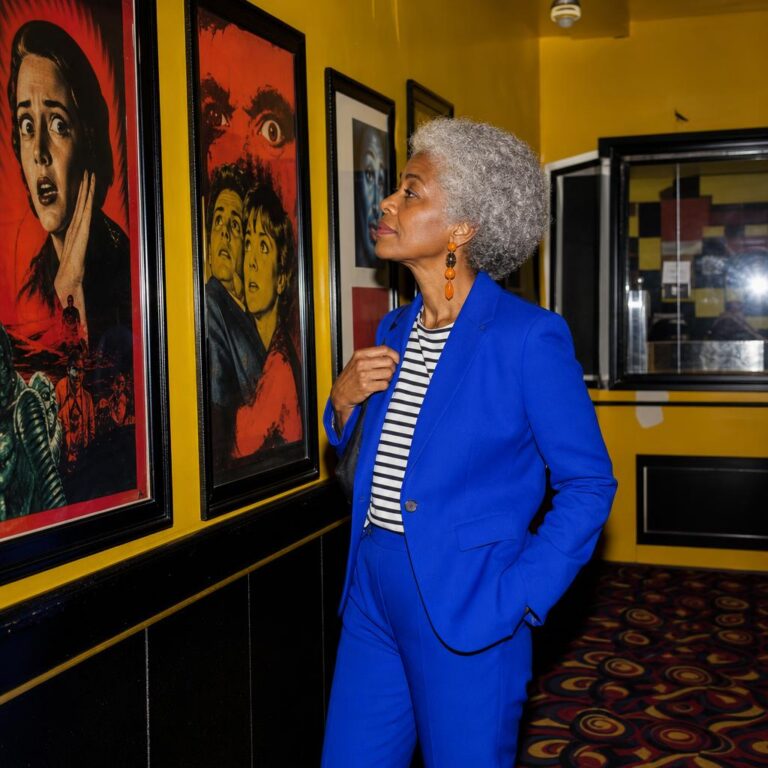 Fictional silver-haired Black woman in a cobalt suit examining colorful movie posters in a yellow cinema lobby.