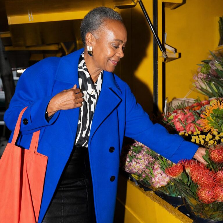 Fictional older Black woman in a cobalt coat choosing flowers at a saturated yellow street-market stall.